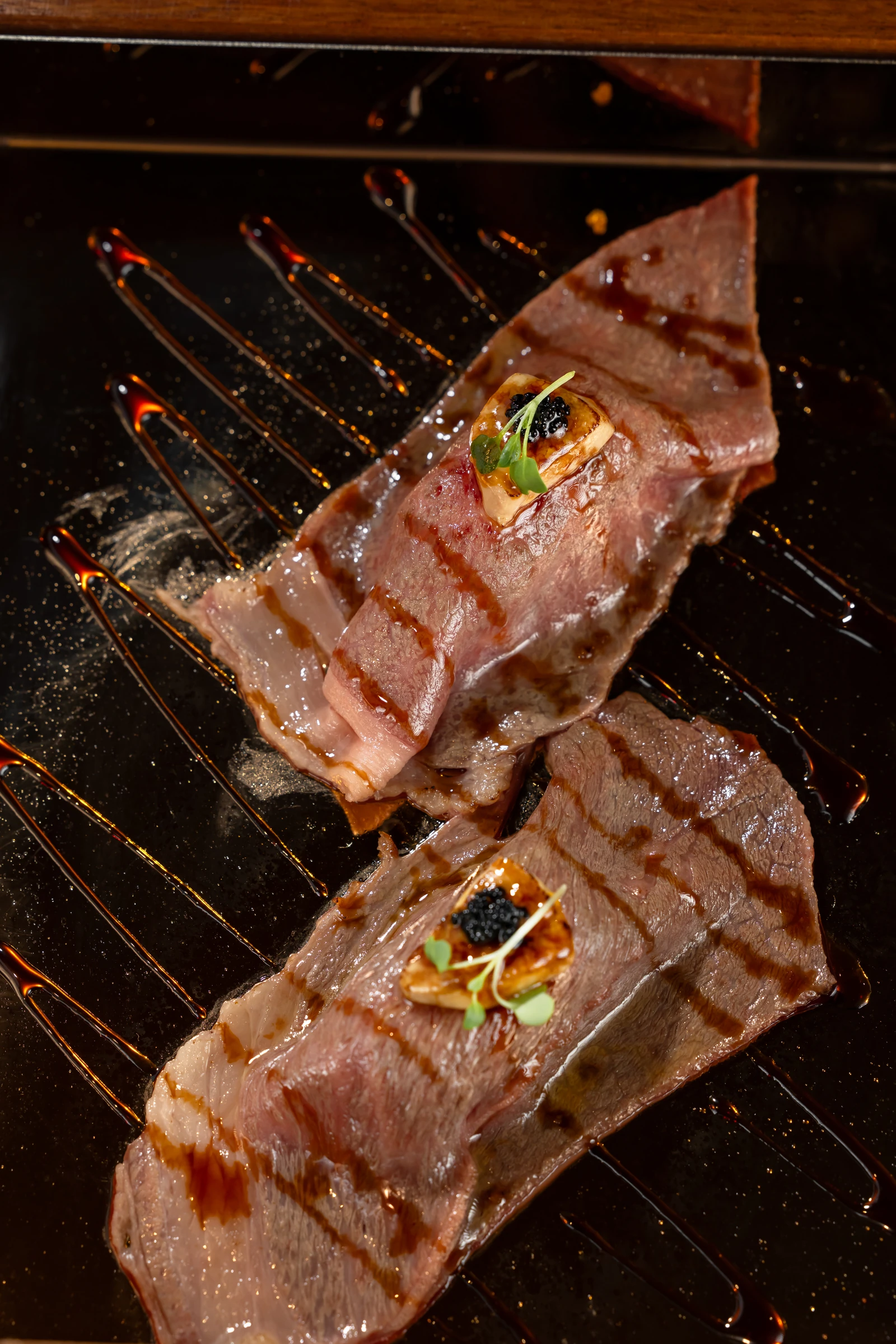 Two seared Wagyu pieces with small garnishes and sauce on a black platter