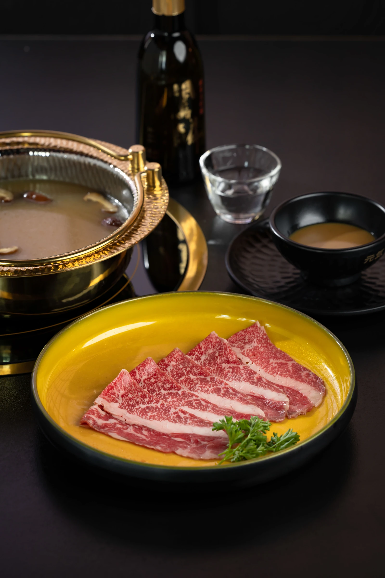 Wagyu slices arranged on a gold-toned plate beside broth and tableware